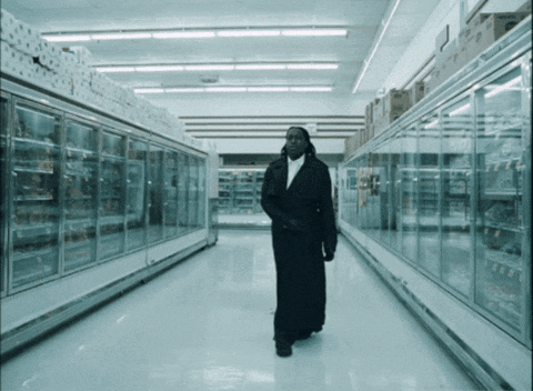 A Man In A Long Black Coat Walks Through An Empty Supermarket Aisle GIF