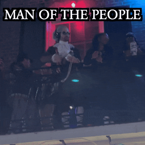 A Man In A Man Of The People Costume Stands In Front Of A Green Light GIF