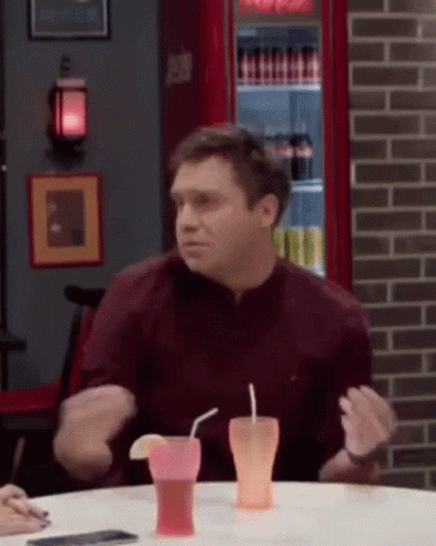 A Man In A Maroon Shirt Is Sitting At A Table With Two Drinks In Front Of Him . GIF