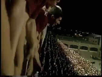 A Man In A Maroon Shirt Is Urinating In Front Of A Crowd Of People . GIF