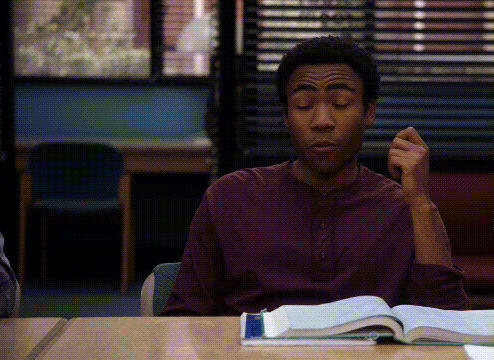 A Man In A Maroon Shirt Sits At A Desk With A Book GIF