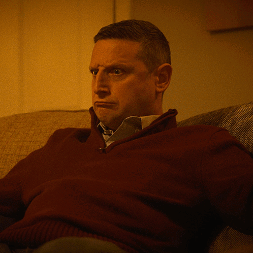 A Man In A Maroon Sweater And Tie Sits On A Couch GIF