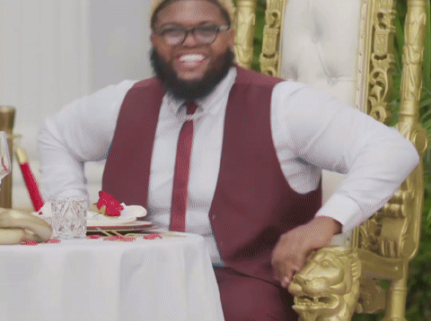 A Man In A Maroon Vest And Tie Is Sitting In A Chair GIF