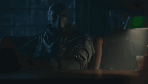 A Man In A Mask Sits At A Desk With A Lamp GIF