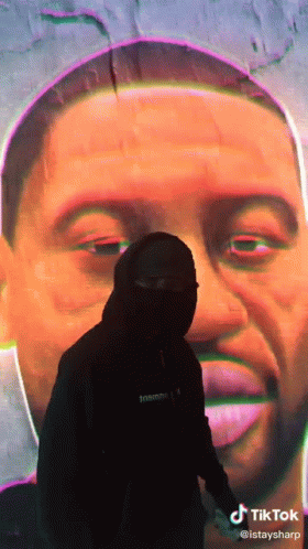 A Man In A Mask Stands In Front Of A Large Painting Of A Man 'S Face GIF