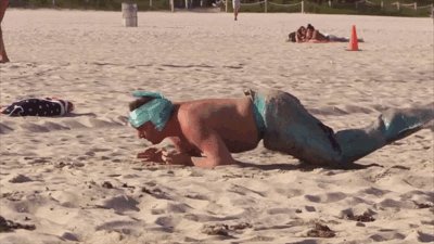 A Man In A Mermaid Costume Is Doing Push Ups On A Sandy Beach GIF