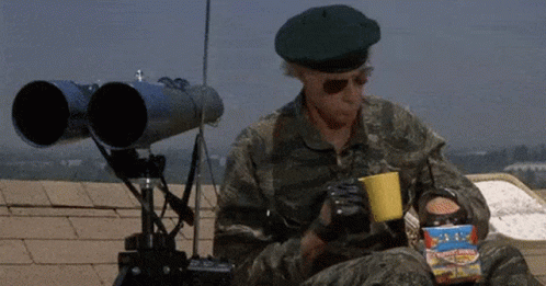 A Man In A Military Uniform Is Sitting On A Roof Holding A Cup Of Coffee And A Box Of Popcorn . GIF