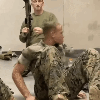 A Man In A Military Uniform Is Sitting On The Floor While Another Man Holds A Rifle . GIF