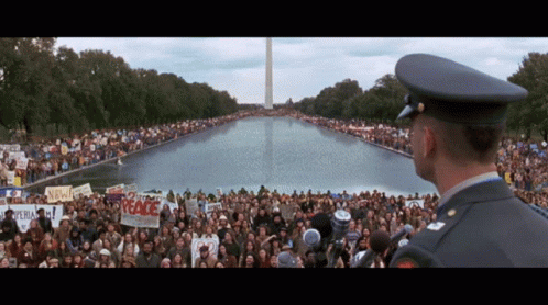 A Man In A Military Uniform Stands In Front Of A Crowd Holding Signs Including One That Says " Peace " GIF