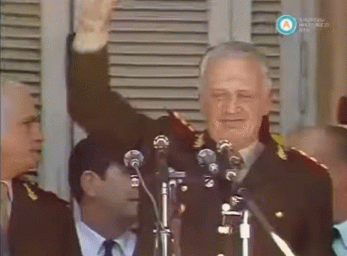 A Man In A Military Uniform Stands In Front Of Two Microphones And Waves His Hand GIF