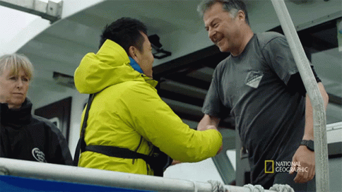 A Man In A National Geographic Shirt Shakes Hands With Another Man On A Boat GIF