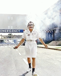 A Man In A Nurse Costume Is Walking Down A Street In Front Of An Emergency Sign GIF