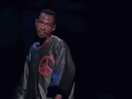 A Man In A Peace Sign Shirt Is Standing In Front Of A Microphone With The Words Next Time Above Him GIF