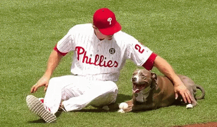 A Man In A Phillies Jersey Sits On The Field With A Dog GIF