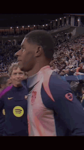 A Man In A Pink And Blue Jersey Is Standing In Front Of A Crowd Of People . GIF