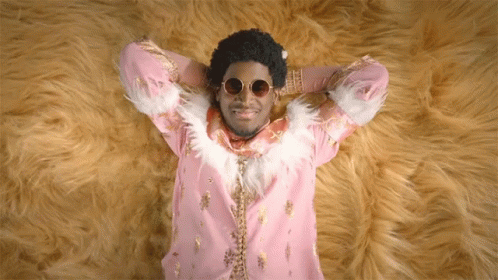 A Man In A Pink And Gold Outfit Is Laying On A Furry Rug GIF