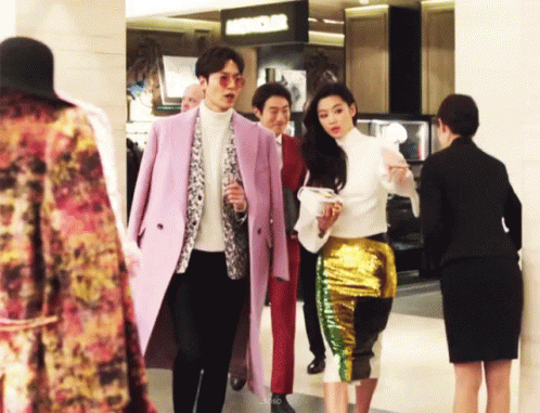 A Man In A Pink Coat And A Woman In A Gold Skirt Are Walking In A Store GIF
