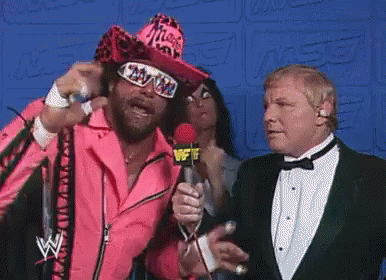 A Man In A Pink Jacket Is Talking Into A Microphone While Another Man In A Tuxedo Watches . GIF
