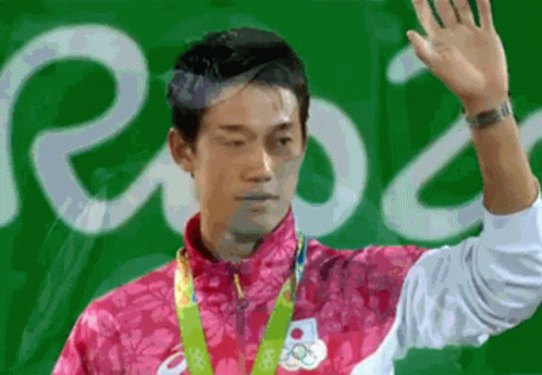 A Man In A Pink Jacket With A Medal Around His Neck Is Waving His Hand In Front Of A Sign That Says Rio GIF