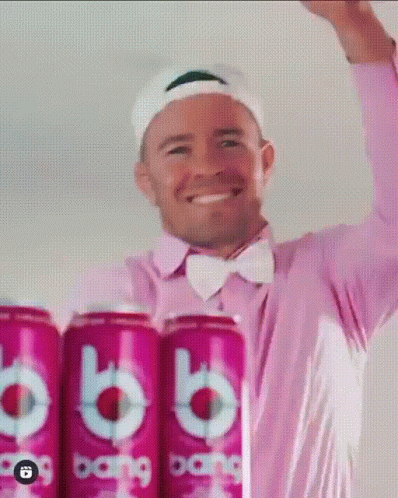 A Man In A Pink Shirt And Bow Tie Is Standing Next To A Row Of Cans Of Bang GIF