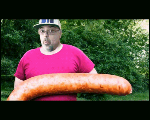 A Man In A Pink Shirt Holds A Very Large Sausage GIF
