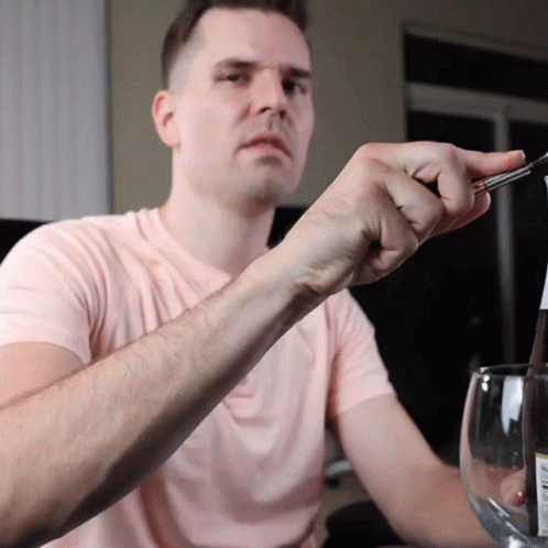 A Man In A Pink Shirt Is Holding A Pair Of Tweezers Over A Bottle GIF