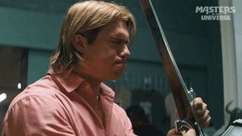 A Man In A Pink Shirt Is Holding A Sword In Front Of A Masters Of The Universe Sign GIF