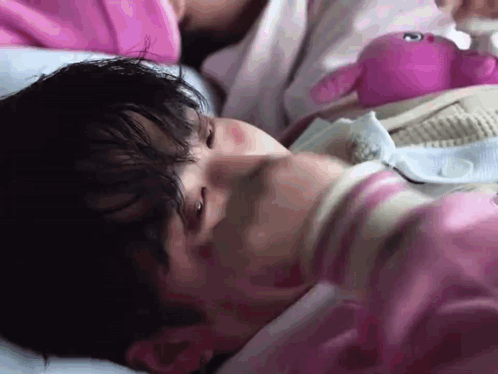 A Man In A Pink Shirt Is Laying In Bed With A Pink Stuffed Animal . GIF