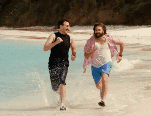 A Man In A Pink Shirt Is Running On The Beach With Another Man GIF