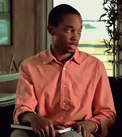 A Man In A Pink Shirt Is Sitting In Front Of A Window Holding A Book GIF