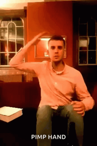 A Man In A Pink Shirt Is Sitting On A Couch And Making A Pimp Hand Gesture . GIF