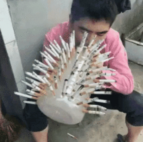 A Man In A Pink Shirt Is Sitting On The Ground Holding A Bowl Of Cigarettes . GIF
