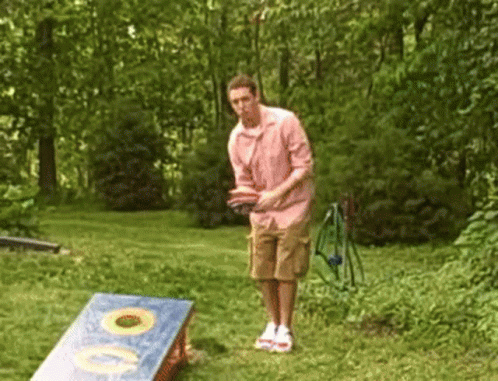 A Man In A Pink Shirt Is Throwing A Frisbee In A Cornhole Board GIF