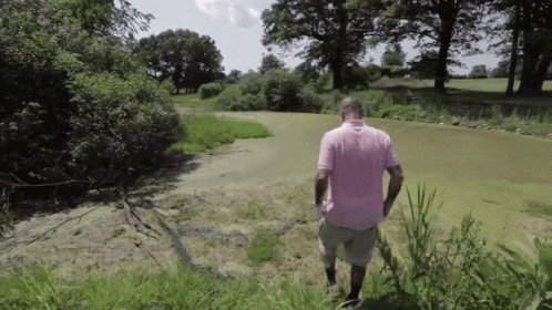 A Man In A Pink Shirt Is Walking Through A Grassy Field GIF