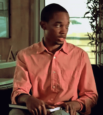 A Man In A Pink Shirt Sits In A Chair Holding A Book GIF