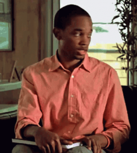A Man In A Pink Shirt Sits In A Chair Holding A Book GIF