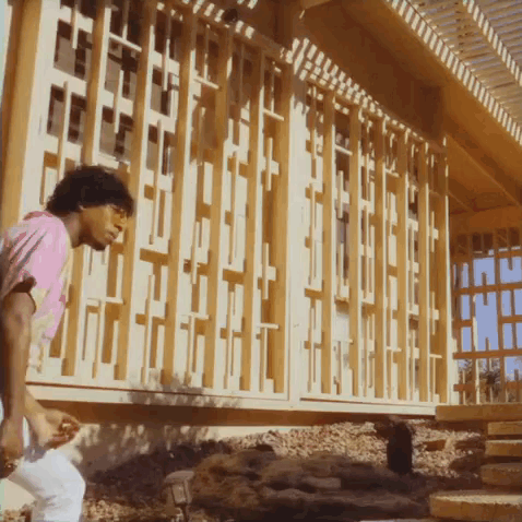 A Man In A Pink Shirt Stands In Front Of A Building With A Wooden Fence GIF
