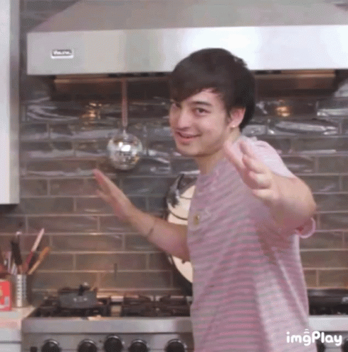 A Man In A Pink Striped Shirt Is Standing In Front Of A Stove With A Ladle Hanging From It .. GIF