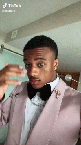 A Man In A Pink Suit And Black Bow Tie Is Taking A Selfie . GIF