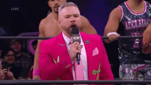 A Man In A Pink Suit Is Speaking Into A Microphone With A Aew Logo GIF