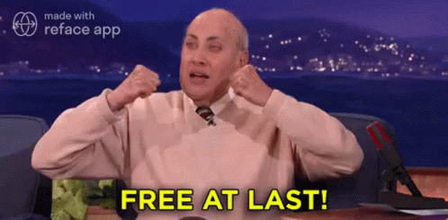 A Man In A Pink Sweater And Tie Is Sitting At A Desk With His Arms In The Air And Says Free At Last . GIF