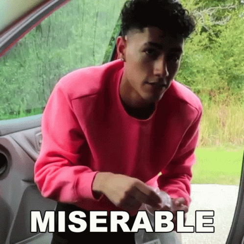 A Man In A Pink Sweater Is Standing In The Doorway Of A Car With The Word Miserable Written On The Side . GIF