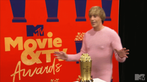 A Man In A Pink Sweater Stands In Front Of A Movie And Tv Awards Sign GIF