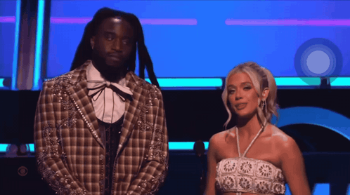 A Man In A Plaid Jacket Stands Next To A Woman In A White Crop Top GIF
