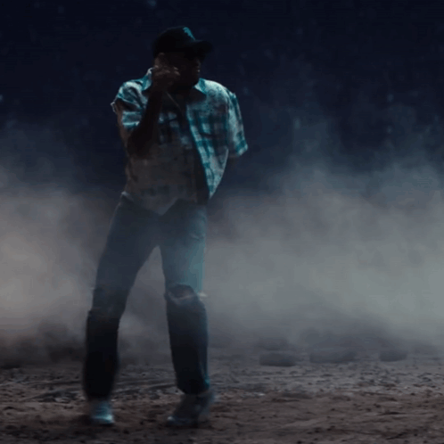A Man In A Plaid Shirt And Cowboy Hat Is Standing In A Dark Room GIF