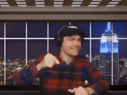 A Man In A Plaid Shirt And Headphones Is Raising His Arms In The Air In Front Of A Large Screen . GIF