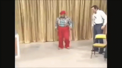 A Man In A Plaid Shirt And Red Pants Stands Next To A Clown GIF