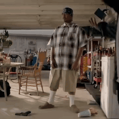 A Man In A Plaid Shirt And Shorts Is Standing In A Garage GIF