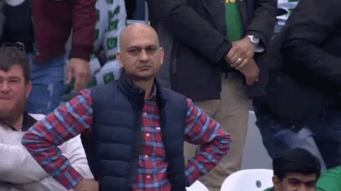 A Man In A Plaid Shirt And Vest Is Sitting In A Crowd Of People With His Hands On His Hips . GIF
