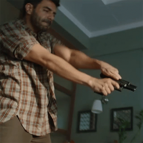 A Man In A Plaid Shirt Holds A Gun In His Hand GIF